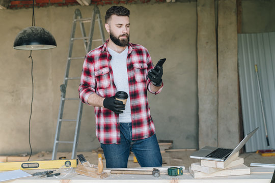 Young bearded businessman, builder, repairman, carpenter, architect, designer dressed in plaid shirt, goggles and gloves, stands in workshop, using smartphone and holds cup of coffee in his hand.