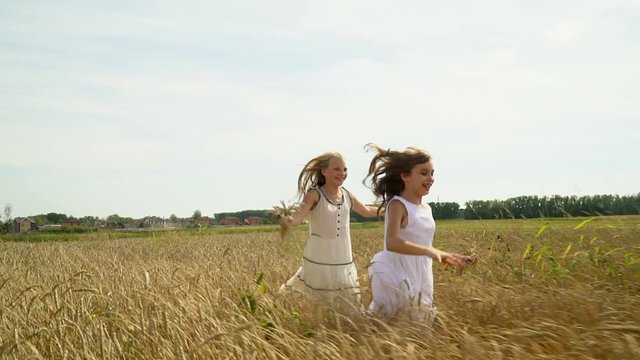 Two girls run on the gold field of wheat. Two beautiful lovely girls in white light dresses run across the field of wheat with ears in hands. Slow motion
