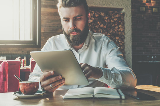 Front view.Young bearded businessman sits in cafe at table,uses digital tablet.On desk is notebook,cup of coffee.Man working,studying.Online education,marketing.E-learning,e-commerce. Instagram filter