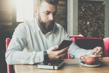 Fototapeta premium Young bearded businessman sits in cafe,office at table,reads information on smartphone screen, drinks coffee.On desk is closed notebook, pen. Online marketing, education, e-learning. Instagram filter.