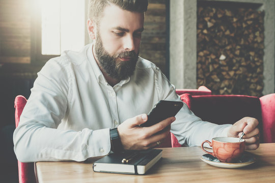 Young bearded businessman sits in cafe,office at table,reads information on smartphone screen, drinks coffee.On desk is closed notebook, pen. Online marketing, education, e-learning. Instagram filter.