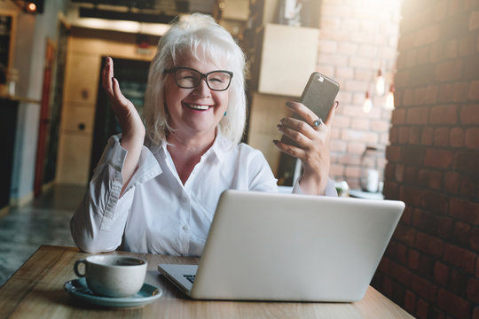 Happy businesswoman sitting at table in front of laptop, holding hands up and smiling, working, learning. In hands of women in a smartphone. Good news. Education for retired people. Online marketing.