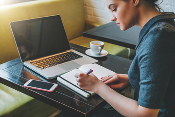 Side view. Young businesswoman sitting at table in cafe in front of laptop, computer and making...