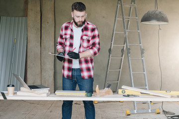 Hipster man is carpenter, builder, designer stands in workshop, holds clipboard and takes notes. On desk is laptop and construction tools, in background stepladder. Repair, construction, carpentry.