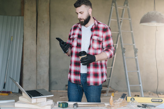 Man is carpenter,builder,designer stands in workshop, holds cup of coffee and uses smartphone. On desk is laptop and construction tools, in background a stepladder. Repair, construction, carpentry.