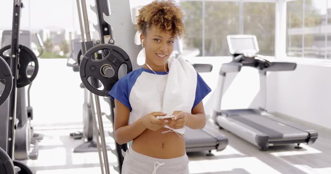 Young attractive woman in sportswear smiling at camera flirty while holding smartphone with headphones on background of gym.