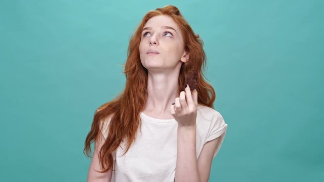 Pleased ginger woman in t-shirt eating cookie and looking at the camera over turquoise background