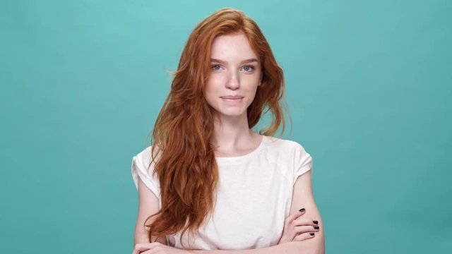 Pleased shy ginger woman in t-shirt posing with crossed arms and looking at the camera over turquoise background