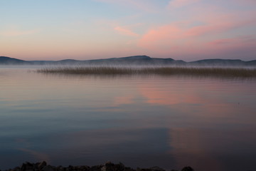 Туманный, ранний, розовый рассвет на озере с горами на заднем плане.