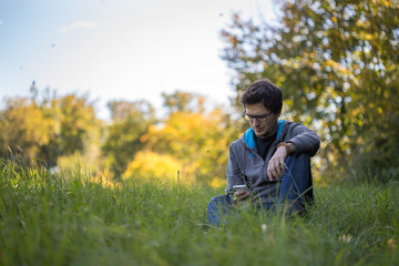 Naklejka na ściany i meble Junger Mann sitzt im Gras, Blick aufs Smartphone, Textfreiraum
