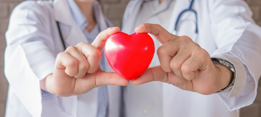 The acting setup of medical work. The Asian young male and female medical doctors carry red heart shape model in their hands in the concept of care your heart. Selective focused