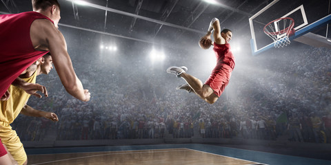 Basketball player makes slam dunk on big professional arena. Player flies through the air with the ball. Opponents try to prevent the ball from hitting the basketball ring. © Alex