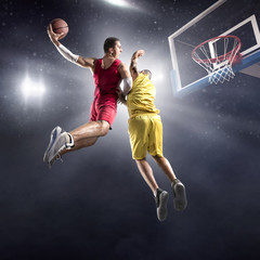 Basketball player makes slam dunk on big professional arena. Player flies through the air with the ball. Opponents try to prevent the ball from hitting the basketball ring.