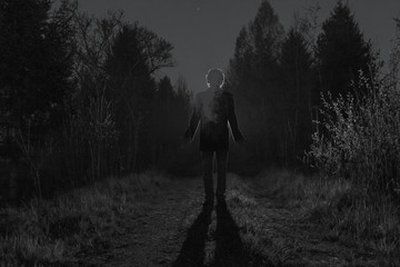 Male silhouette at the dark forest pathway through the bushes in the night. Man standing on the road against the car headlights. Mystery ghost concept. Film grain filter, soft focus. Black and white.