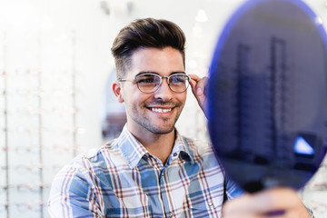 Handsome young man choosing eyeglasses frame in optical store.