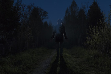 Male silhouette at the dark forest pathway through the bushes in the night. Man standing on the road against the car headlights. Mystery ghost concept. Film grain filter, soft focus.