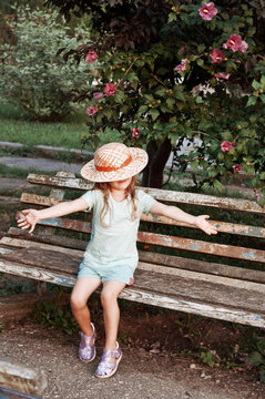 Little girl in a hat sitting on an old bench. Bush with pink flowers. The girl's face is closed hat.