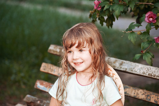 Portrait of a little girl. The girl is sitting on an old bench. Bush with pink flowers.