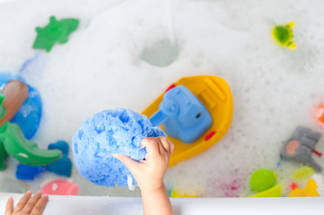 Hands of a toddler holding blue sponge with bathtub on the background
