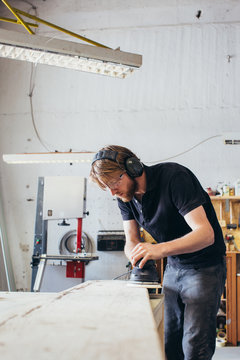 Young Blond Carpenter Sanding Wood Plank With Random Orbital Sander in Bright Workshop