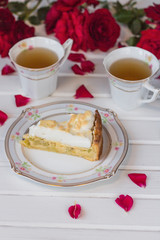 A piece of pie with rhubarb and meringue  is on a white table near red roses and cups of tea.