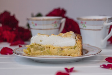 A piece of pie with rhubarb and meringue  is on a white table near red roses and cups of tea up close.
