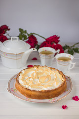 Meringue pie on a white table near teapot and cups and red roses.