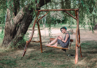 Young beautiful girl sits in a park on a wooden swing made of eco-friendly materials, resting on the day off
