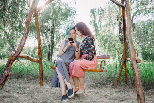 Two girls stits on wooden swing, looking at smartphone. Browsing internet, posting photos in social media