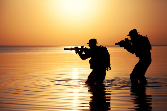 Army soldiers with rifles orange sunset silhouette in action during raid crossing river in the water