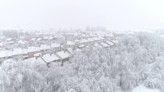 AERIAL: Flying over quiet suburban town with roofs covered in fresh snow on winter day. Idyllic row houses on snowy winter day. Flying above suburb rooftops and snowy backyards in peaceful suburbia.