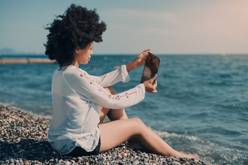 Rear view of mixed girl with afro hair taking pictures of waterscape via digital pad; black young lady in wet chemise sitting on peeble shoreline and photographing sea water using her digital tablet - Powered by Adobe