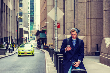 American man traveling in New York, wearing blue long overcoat, scarf, cuffed knit beanie hat with pom pom, carrying back bag, sitting on vintage street, texting on cell phone. Car on background..