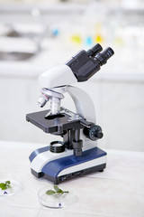 Close-up shot of modern binocular microscope standing on desk of chemistry lab, seedlings lying in Petri dishes prepared for carrying out experiment, blurred background