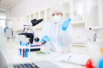 Concentrated female researcher wearing hazmat suit filling test tube with pipette while  working on...