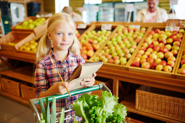 Little buyer with shopping list pushing cart with fresh vegetables