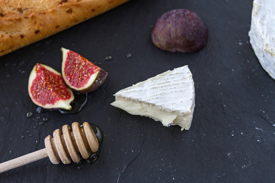 Triangular appetizing piece of camembert cheese and cheese head, pieces of figs in honey and a wooden spoon for honey and baguette on a graphite black background.