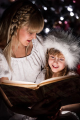Mother reading her daughter a book at night under the Christmas tree