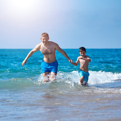 Dad and son are running in the water along the sea shore. They are holding hands and moving towards the camera.