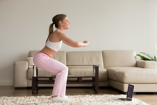 Young sporty woman doing two-leg deep squat morning exercise standing in living room, teen girl burning fat calories with online fitness training help crouching for buttocks lift at home, side view