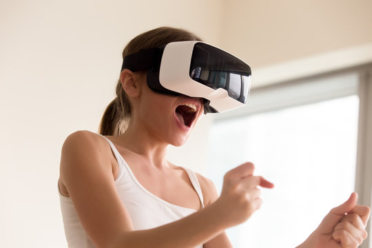 Young female gamer in wearable VR headset playing action 3d video game at home, excited teen holding wheel and screaming driving digital racing car in simulator application, virtual gaming concept