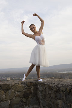 young ballerina in white dress and satin ballet shoes posing on the edge of old fortress wall on a grey sky background