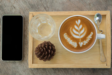  cup of coffee on wooden tray with pine cones and water and smartphone on table.