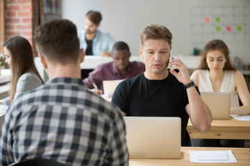 Serious casually dressed businessman talking on cellphone in shared coworking office at work desk in front of laptop. Important deal discussion, business negotiations, client consulting concept.