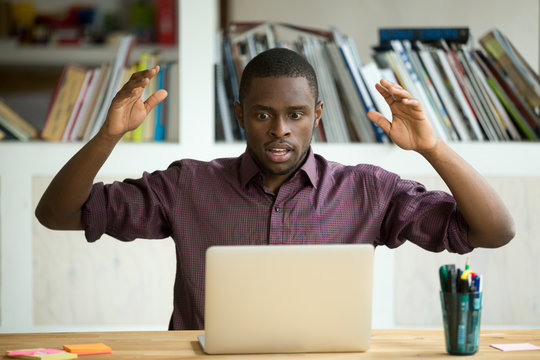 Shocked Young African American Office Worker Looking At Laptop Screen Throwing Arms In The Air. Horrified Small Business Owner Sees App Error, Lost Documents Because Of Unexpected Bad Computer Crash.