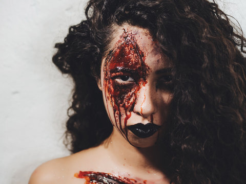 Scary portrait of young zombie girl with Halloween blood makeup. Beautiful latin woman with curly hair looking into camera in studio. Living dead lady.