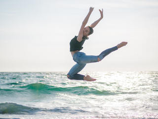 Fototapeta premium Beautiful scene of a dancing ballerina in black top and jeans on sandy beach ocean or sea in morning. Beautiful brunette woman practicing stretching and exercises.Copy space