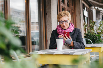 Adult blonde woman browsing smartphone and having drink from paper cup in outside cafe.