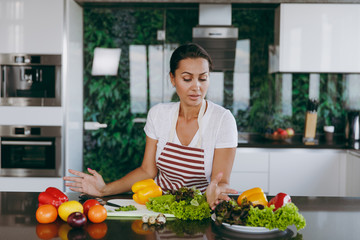 A young confused and thoughtful woman in apron decides what to cook in the kitchen. Healthy food -...