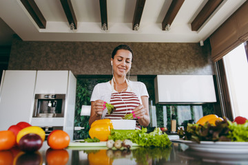 Young woman with headphones in the ears holding vegetables in hands in kitchen with laptop on the table. Vegetable salad. Diet. Dieting concept. Healthy lifestyle. Cooking at home. Prepare food